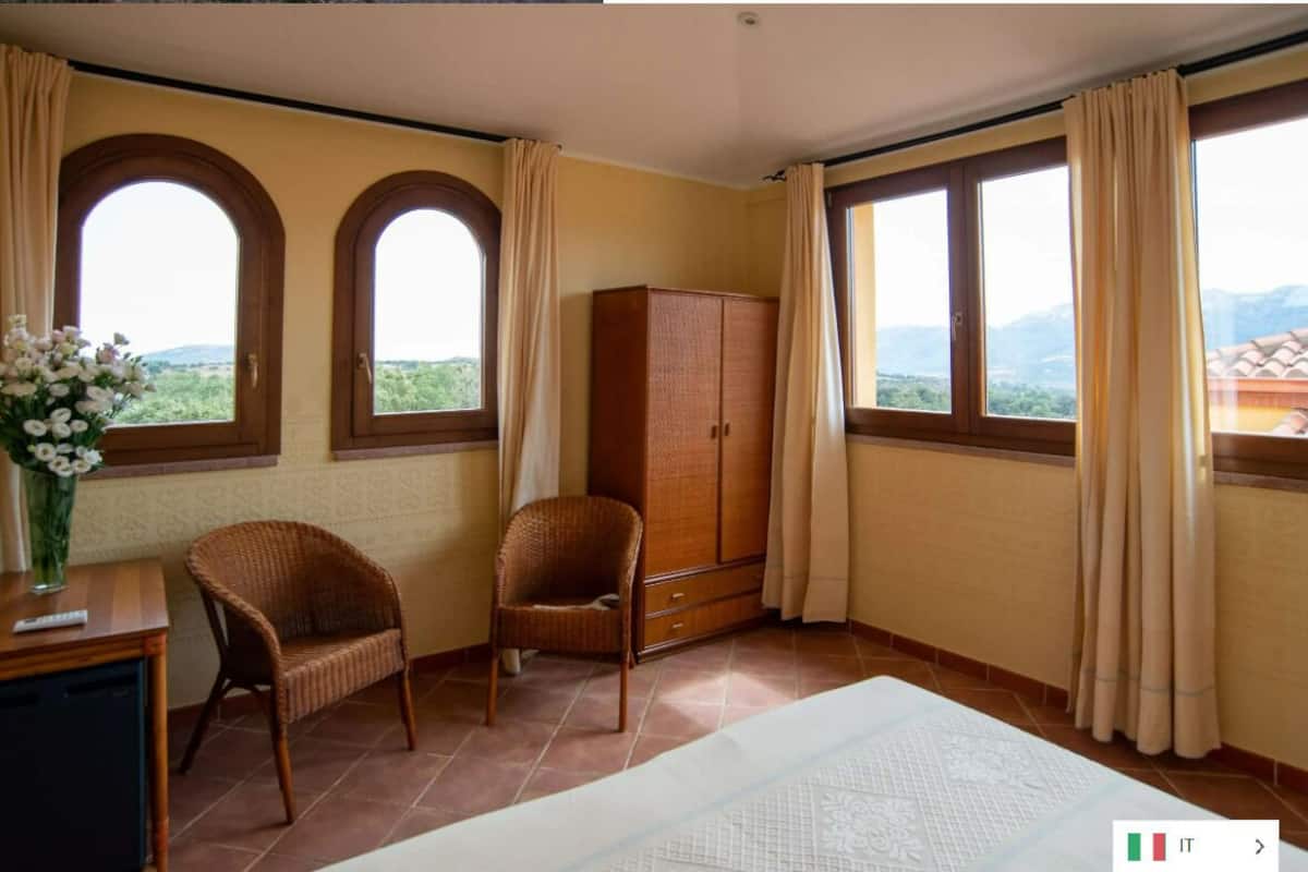 Una habitación con dos ventanas de arco, un armario de madera, una silla de mimbre y una cama.