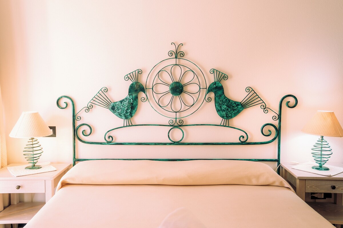 Una cama con cabecera metálica verde que presenta dos pájaros y un diseño circular. Hay dos mesitas de noche con lámparas a cada lado de la cama.