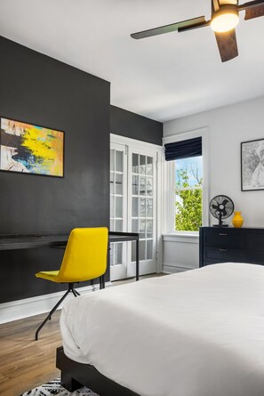 A bedroom with a bed, a yellow chair, a desk, a window with a view, and a ceiling fan.