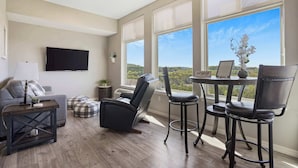 A modern living room with a gray sofa, a black leather recliner, a dining table with two chairs, a flat-screen TV mounted on the wall, and large windows offering a view of the outdoors. - Majestic View Getaway w/ Beautiful Lake Views! (Branson)