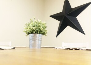 A metal bucket with a plant on a wooden table, a black star decoration on the wall, and white placemats.