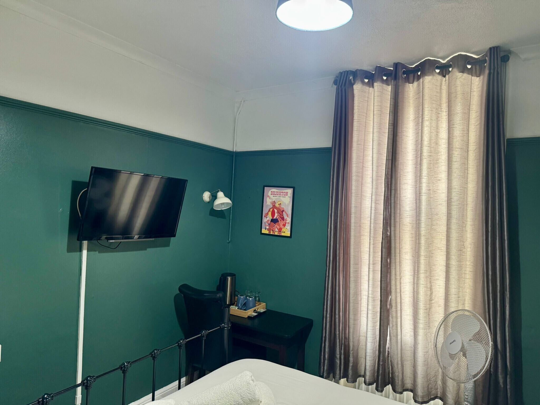 A room with teal walls, a flat-screen TV mounted on the wall, a desk with a chair, a bed, and a window with curtains.