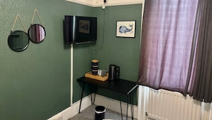 Una habitación pequeña con una pared verde, un televisor negro montado en la pared, un cuadro enmarcado de una ballena, una pequeña mesa de madera con una tetera y tazas, y un cubo de basura negro.