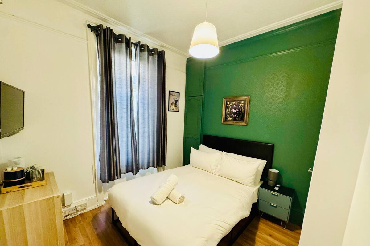 A neatly made bed with white linens and two towels, a bedside table with a lamp, a green accent wall, and a window with curtains.