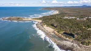 An aerial view of a coastal area with a beach, rocky coastline, and scattered buildings. - Baan Sabai - Oceanstays (Angourie)
