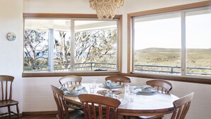 A dining area with a round table set for four, large windows offering a view of trees and hills, and a beaded chandelier hanging above. - Baan Sabai - Oceanstays (Angourie)