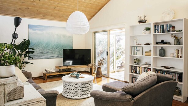 A modern living room with a wooden ceiling, a large flat-screen TV, a white round coffee table, and a bookshelf filled with books and decorative items. - Baan Sabai - Oceanstays (Angourie)
