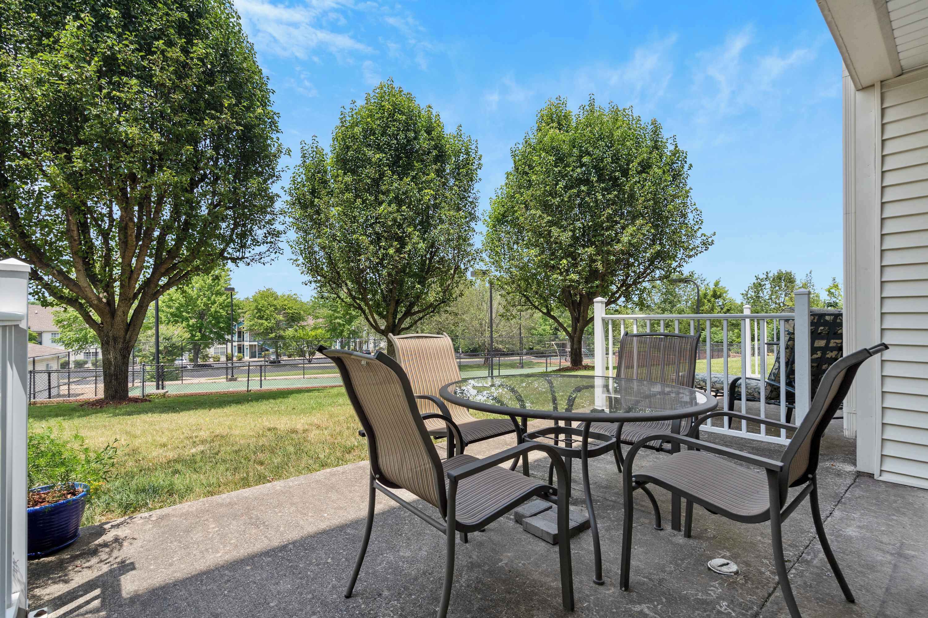 Un patio al aire libre con una mesa redonda y cuatro sillas, rodeado de árboles y vistas a un parque.