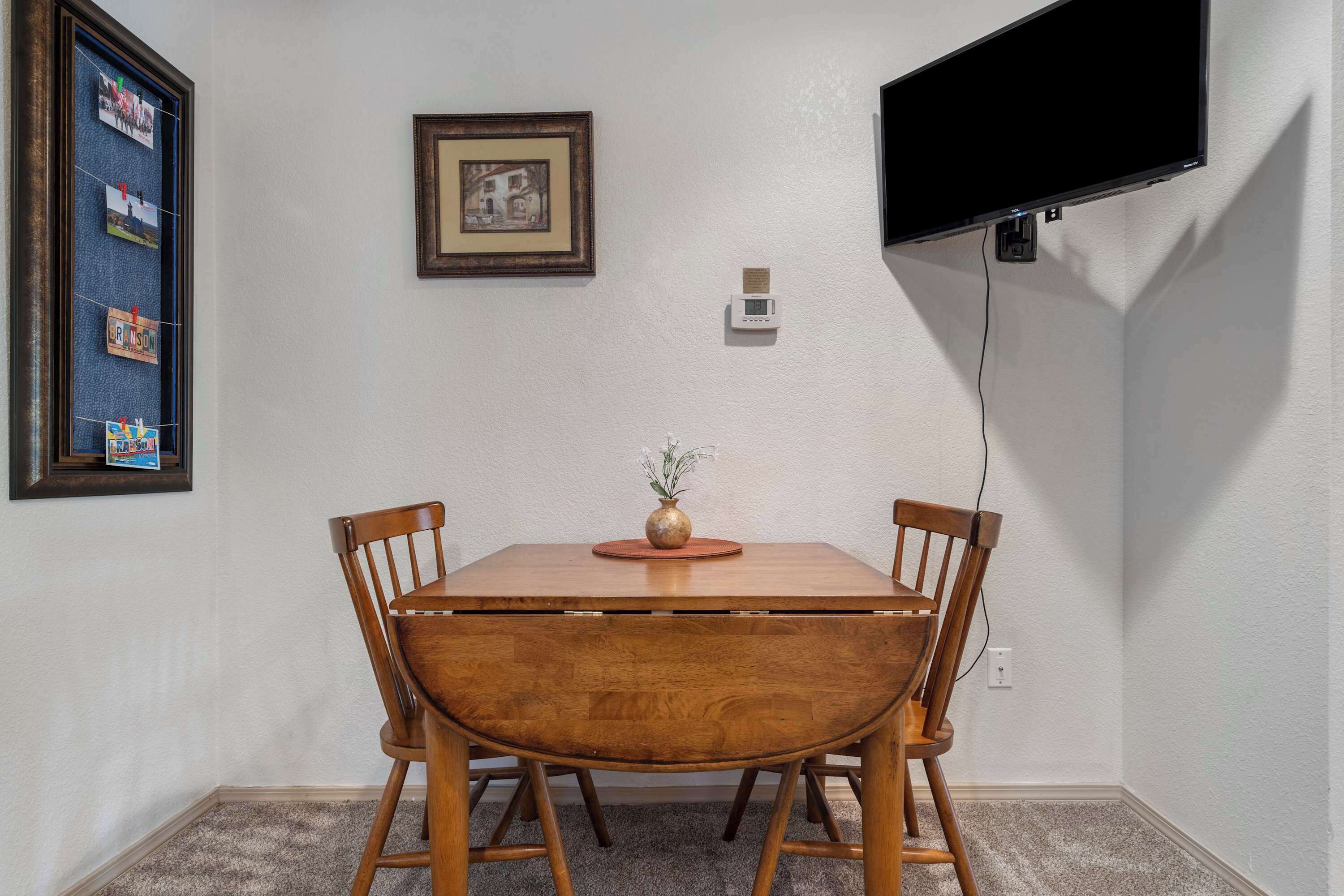 Un coin repas avec une table et des chaises en bois, un tableau encadré, un thermostat et un téléviseur fixé au mur.
