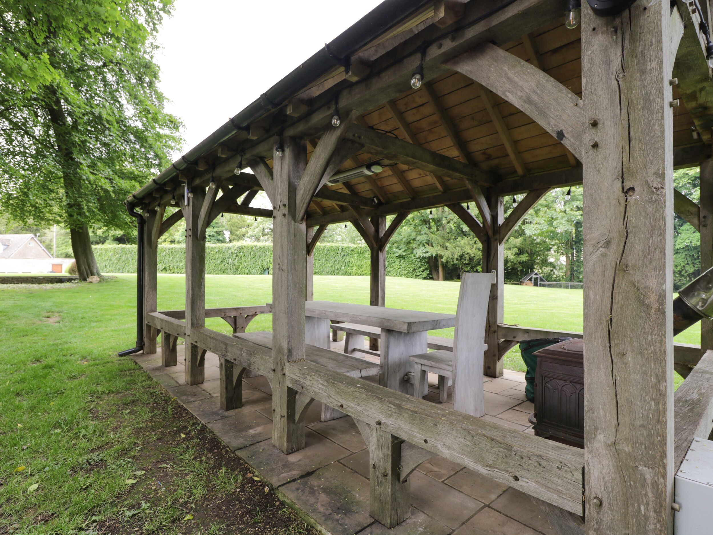 A wooden pavilion with benches and tables, surrounded by a grassy area and trees.