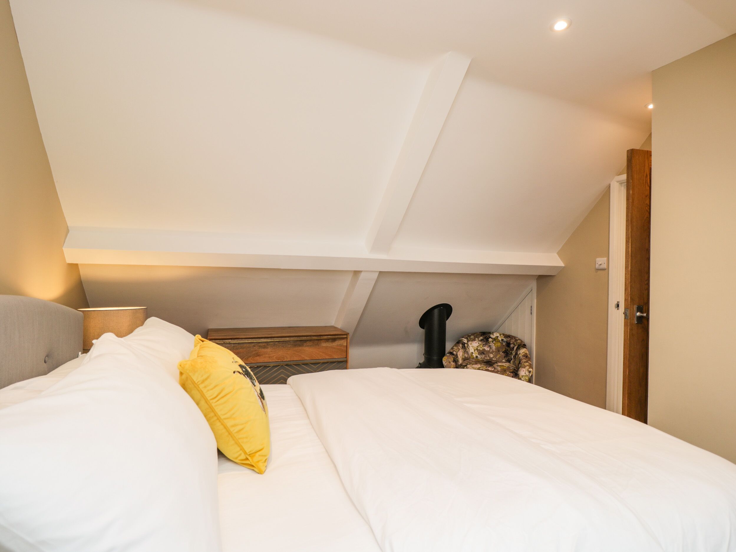 A bedroom with a sloped ceiling, a bed with white bedding and a yellow pillow, a wooden chest, and a black heater.
