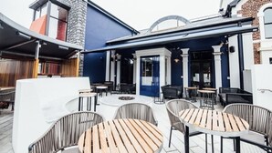 An outdoor patio area with round tables and chairs, a stone wall, and a blue building facade.