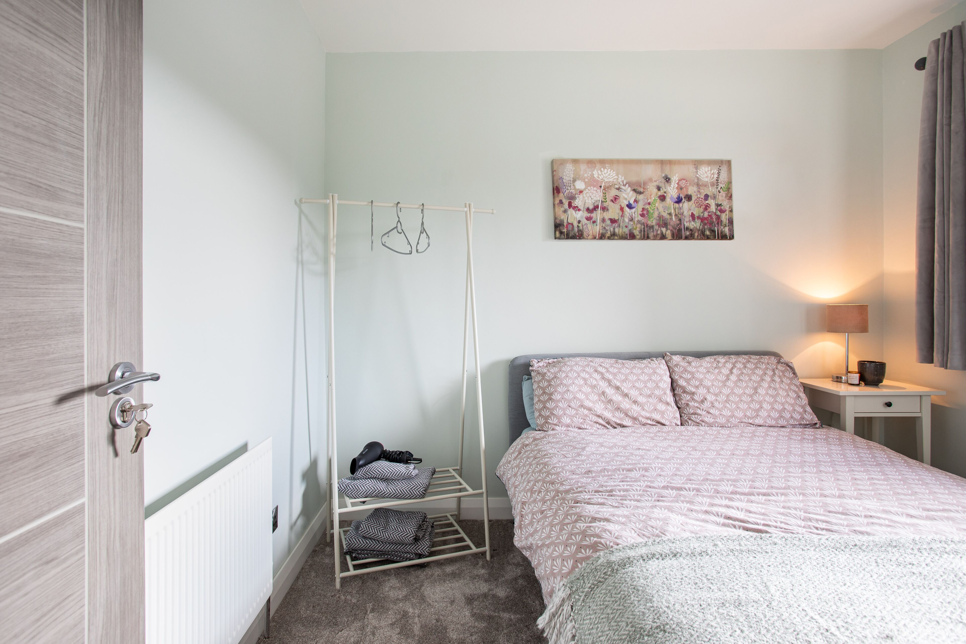 A bedroom with a bed, a radiator, a drying rack, a wall art piece, and a bedside table with a lamp.