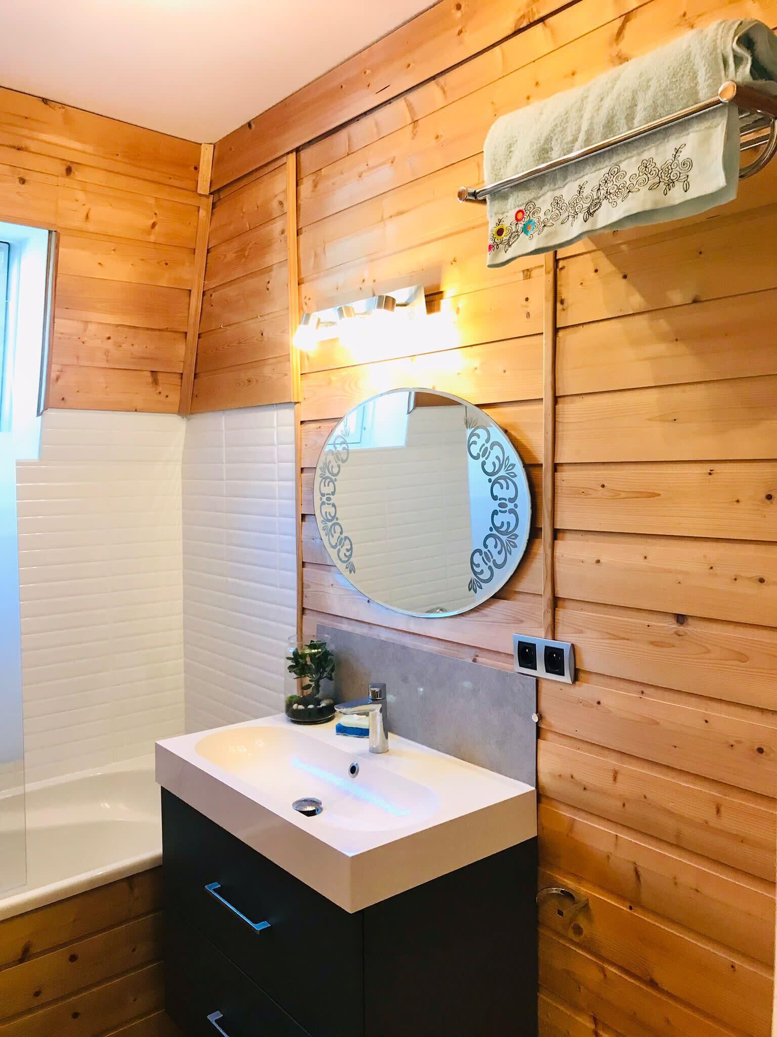 Un bagno con pareti in legno, uno specchio tondo con cornice decorativa, un lavandino bianco con rubinetto azzurro e un porta asciugamani sopra lo specchio.