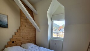 Une chambre à coucher avec une tête de lit en bois, un lit recouvert de linge de maison blanc, une fenêtre donnant sur des maisons et des montagnes, et un escalier en bois.