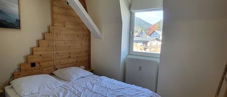 Un dormitorio con cabecera de madera, cama con ropa de cama blanca, ventana con vistas a casas y montañas, y una escalera de madera.