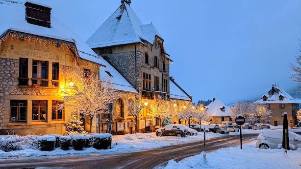 Une rue enneigée bordée d’immeubles, dont un bâtiment en pierre sur lequel on peut lire l’inscription « École communale ».