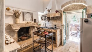 A rustic kitchen with a stone fireplace, wooden bar stools, and a view of the outdoor patio. - Trullo Orto dei Nonni - Le Case di Valentina (Ceglie Messapica)