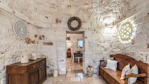A rustic interior with a wooden bench, a white wall with a decorative wreath, and a stone floor. - Trullo Orto dei Nonni - Le Case di Valentina (Ceglie Messapica)