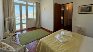 Een hotelkamer met een bed, een groen kleed, een rieten stoel, uitzicht op zee door grote ramen en een schilderij aan de muur.