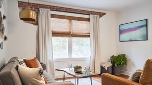 A living room with a sofa, a coffee table with decorative items, a window with curtains, and a painting on the wall. - Hopp's Downtown Loft (McPherson)