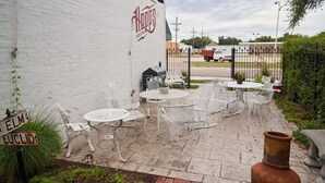 A patio with white metal chairs and tables, a brick pathway, and a white wall with a sign. - Hopp's Downtown Loft (McPherson)