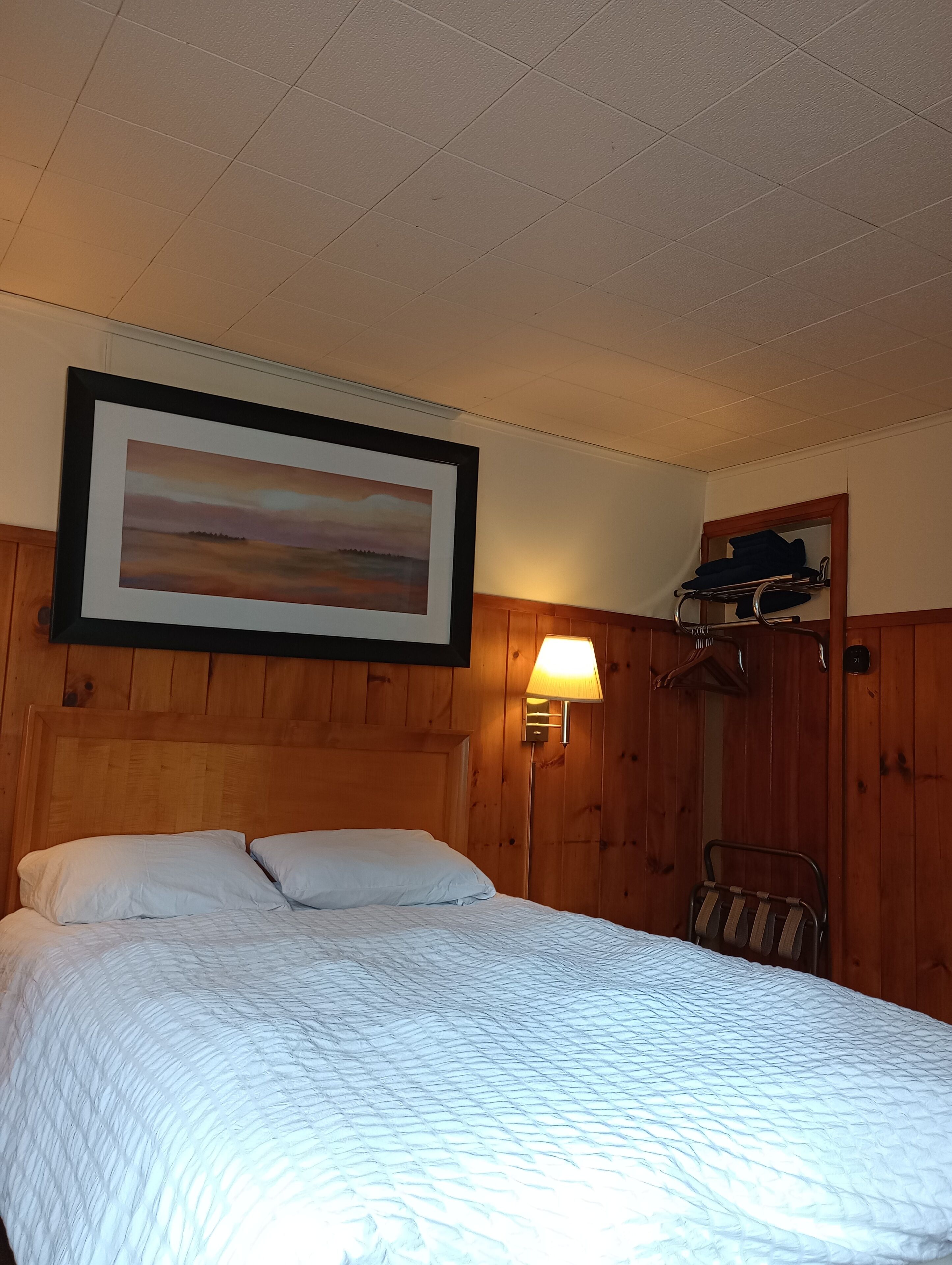 A hotel room with a bed, a framed picture on the wall, a wall-mounted lamp, and a coat rack.