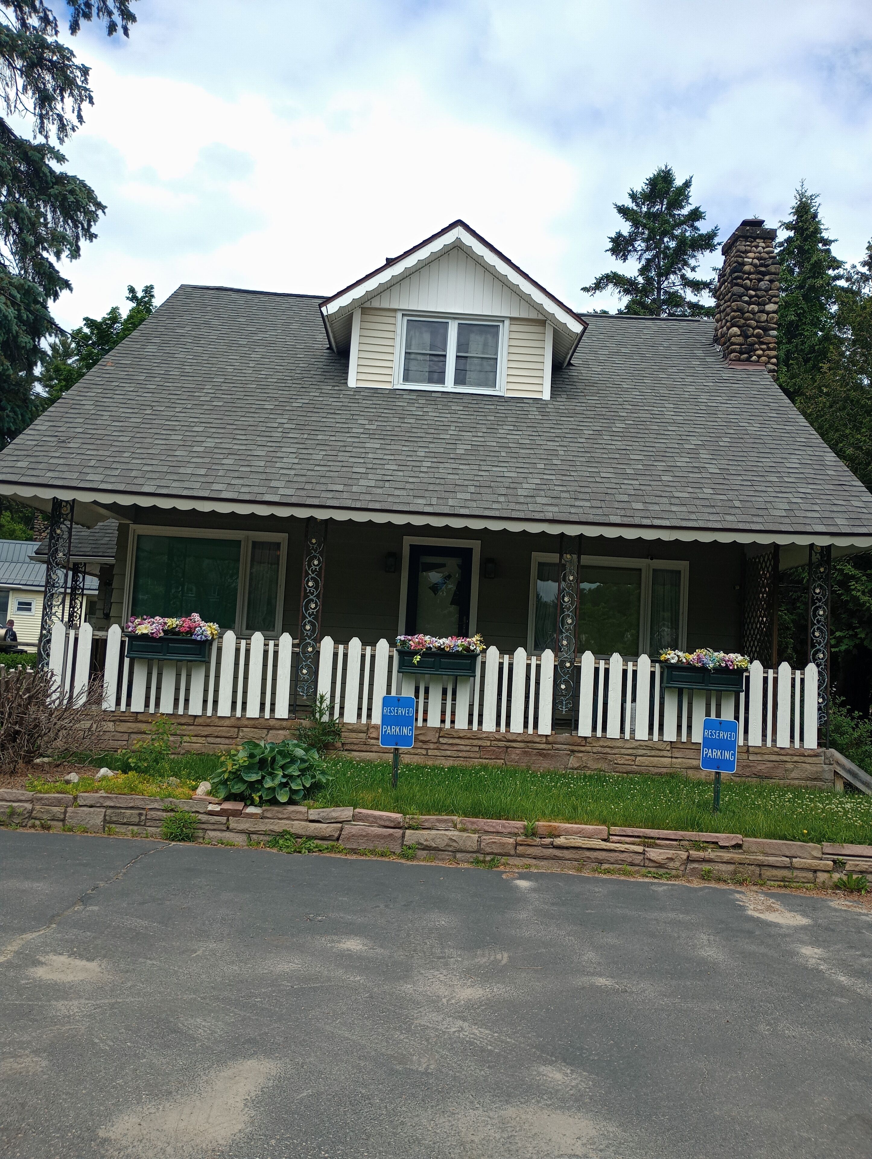 A two-story house with a gabled roof, a front porch with a white picket fence, and two flower boxes. There are two 'RESERVED PARKING' signs in front of the house.