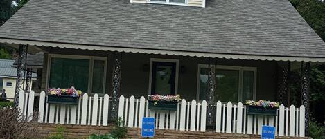 A two-story house with a gabled roof, a front porch with a white picket fence, and two flower boxes. There are two 'RESERVED PARKING' signs in front of the house.