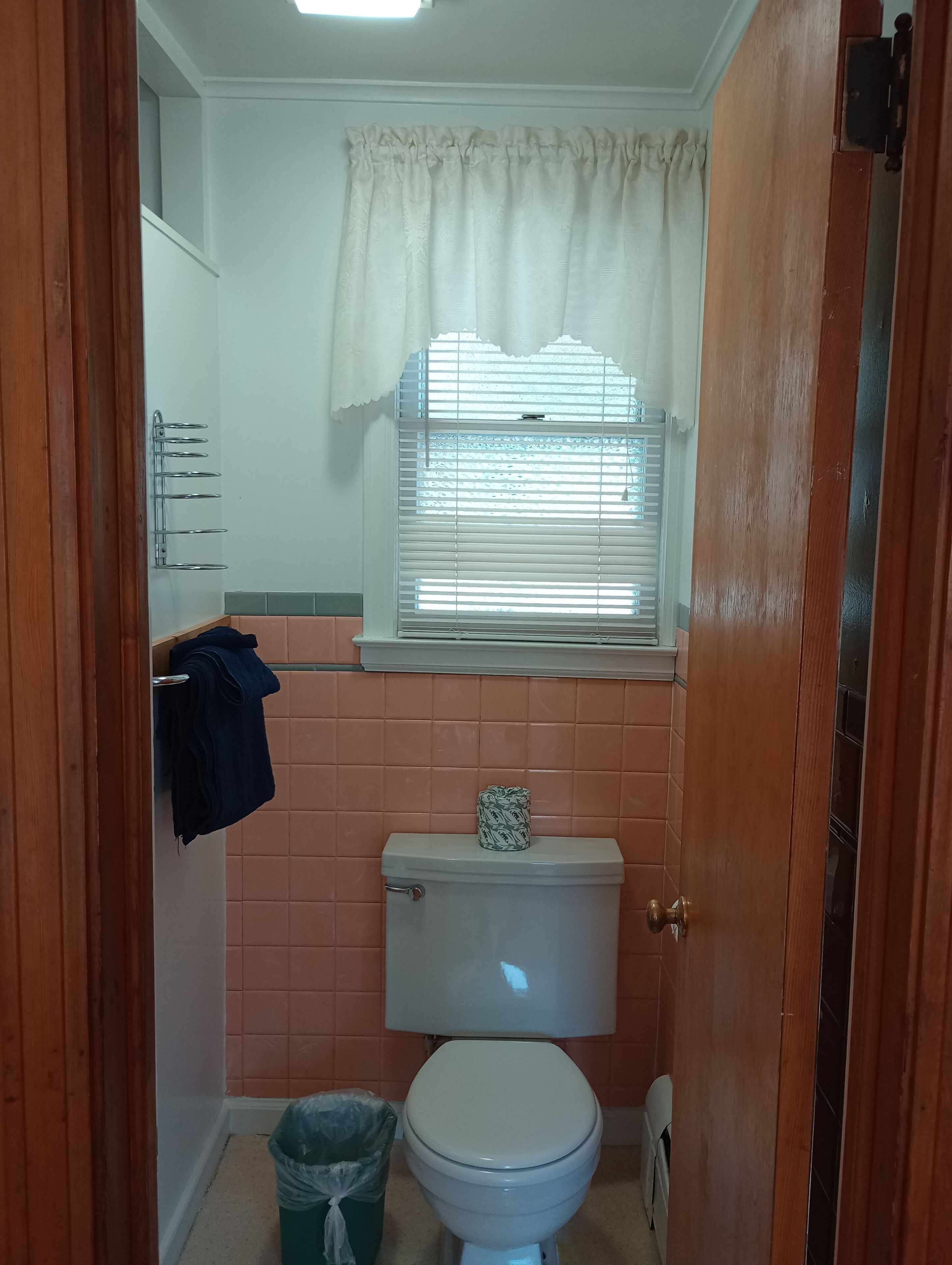 A small bathroom with a toilet, a window with blinds, a towel rack, and a trash bin.