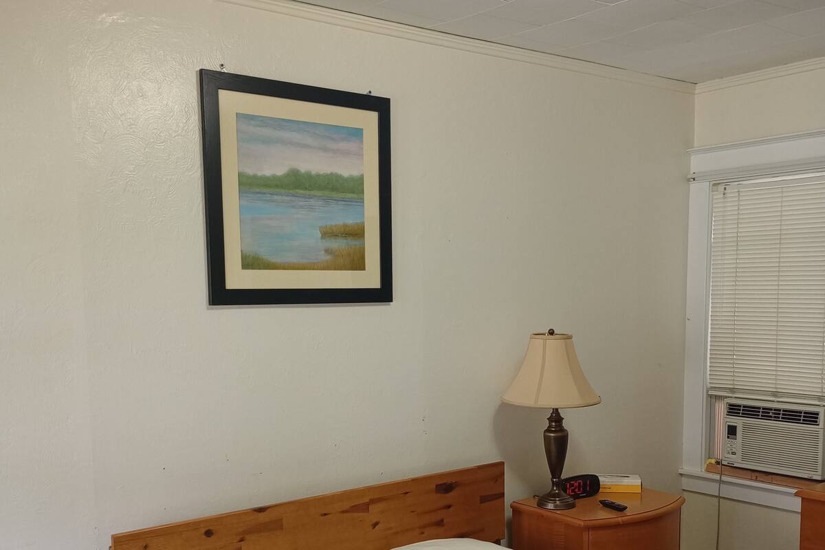 A bedroom with a bed, a nightstand with a lamp, a digital clock, and a window with blinds.