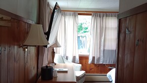 Um quarto com escrivaninha de madeira, luminária, lixeira, cama e janela com cortinas.