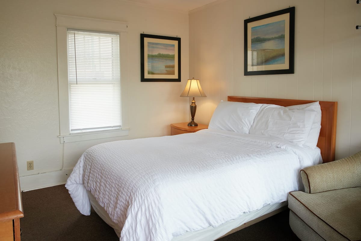 A neatly made bed with white bedding, a wooden headboard, a nightstand with a lamp, and two framed pictures on the wall.