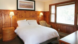 Quarto com parede de painéis de madeira, cama com roupa de cama branca, duas luminárias de cabeceira, uma cadeira e uma janela com persianas.