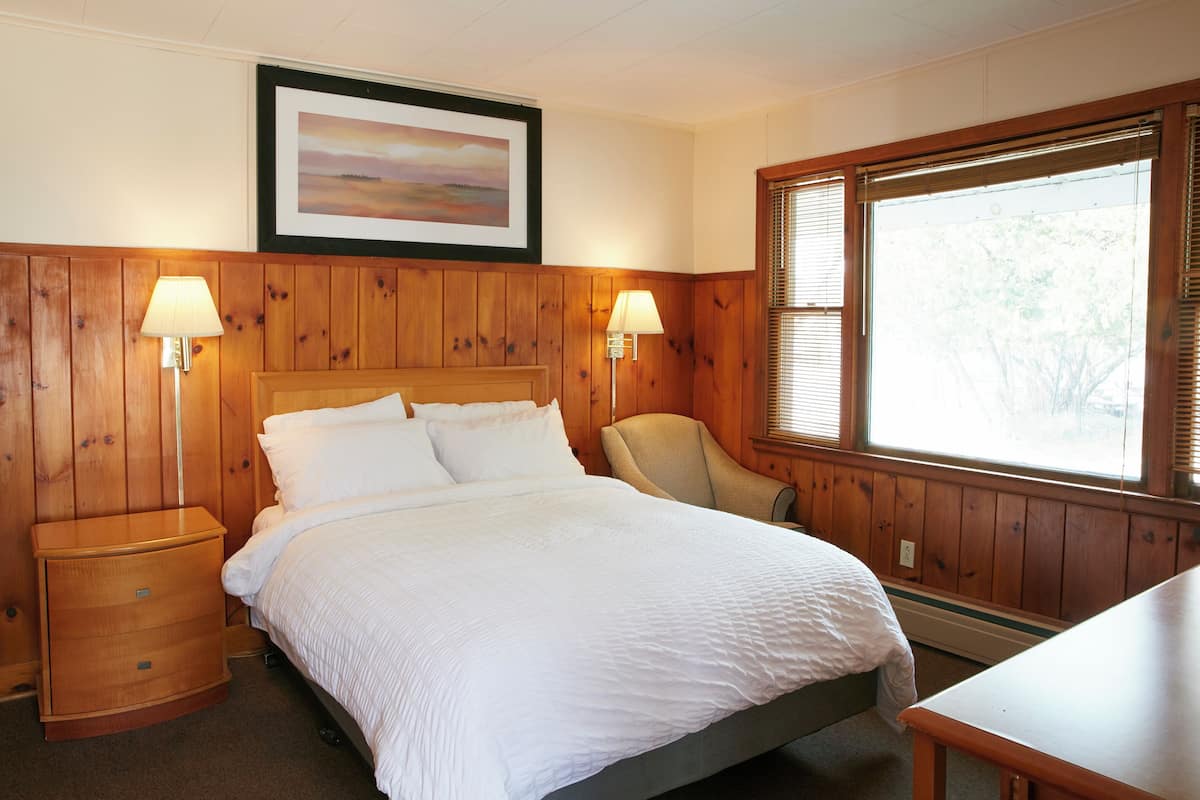 A bedroom with a wooden paneled wall, a bed with white bedding, two bedside lamps, a chair, and a window with blinds.