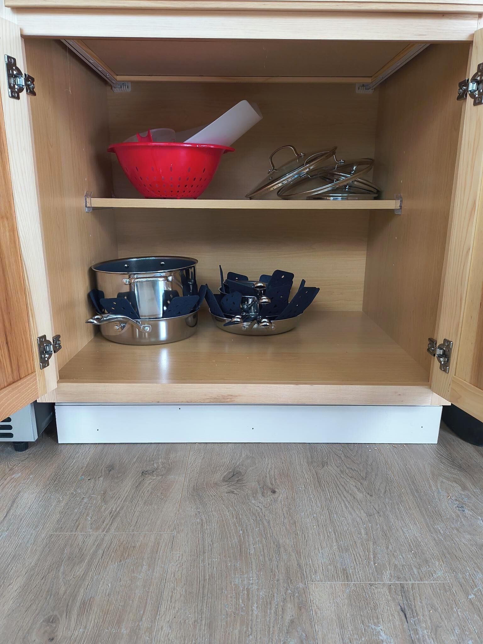 Een houten kast met twee planken; de bovenste plank bevat een rode zeef en een witte plastic bak, terwijl de onderste plank een roestvrijstalen pan en een blauwe zeef bevat.