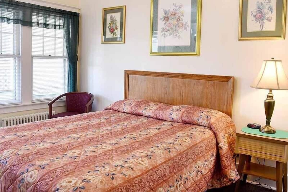 A bedroom with a bed, a chair, a bedside table with a lamp, and two framed floral pictures on the wall.