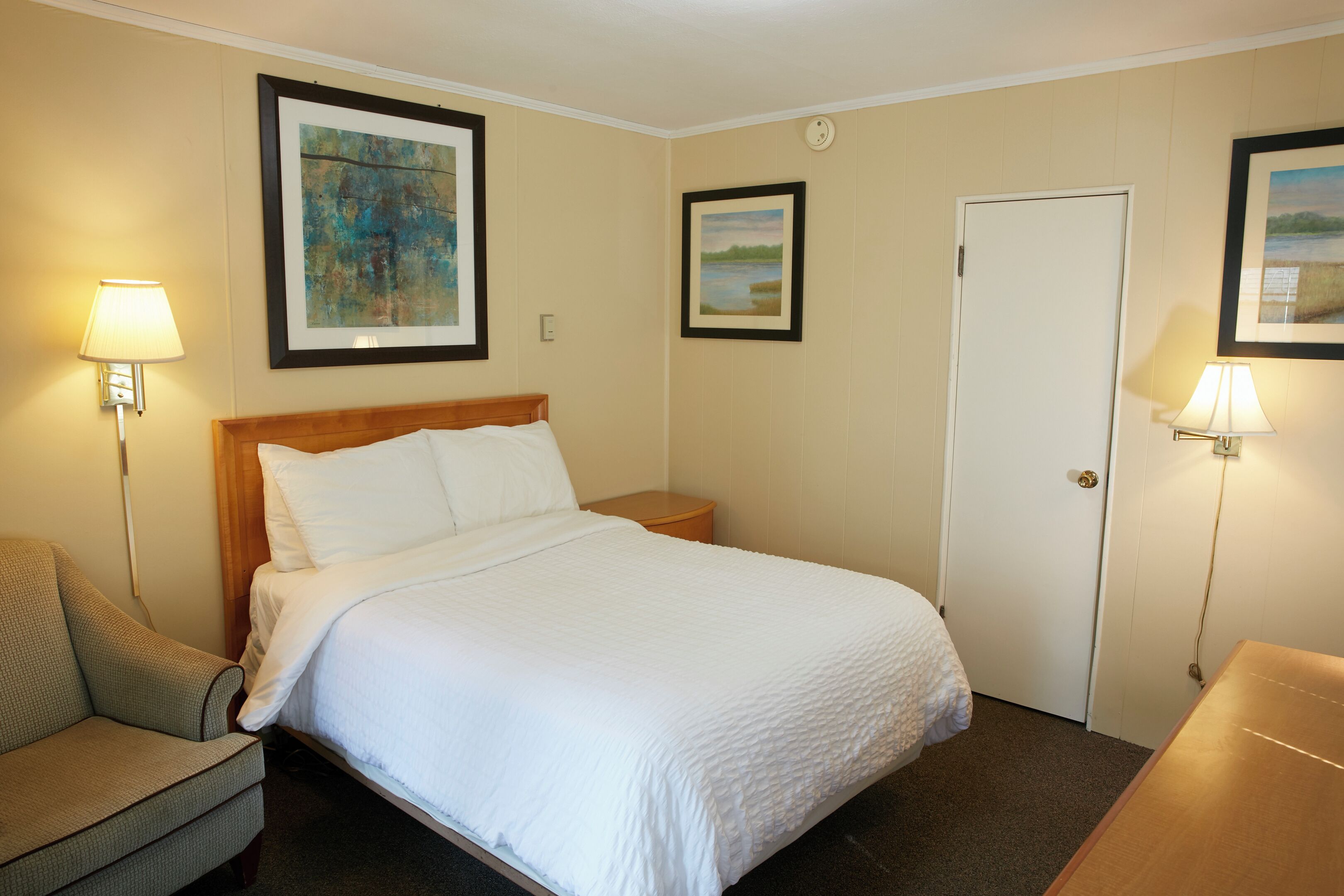 Une chambre d’hôtel comprenant un lit, une chaise, une table de chevet et deux lampes fixées au mur.