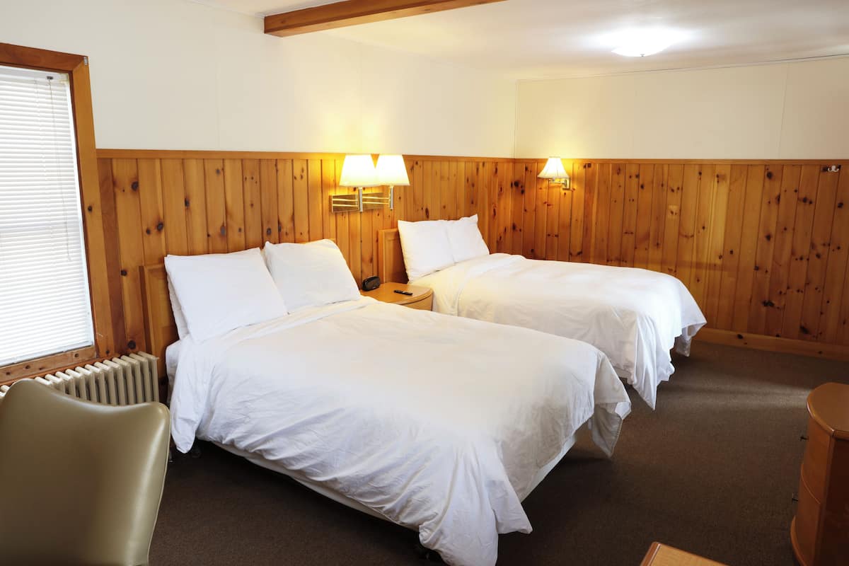 A hotel room with two beds, wooden panel walls, a radiator, a chair, and a wooden side table.