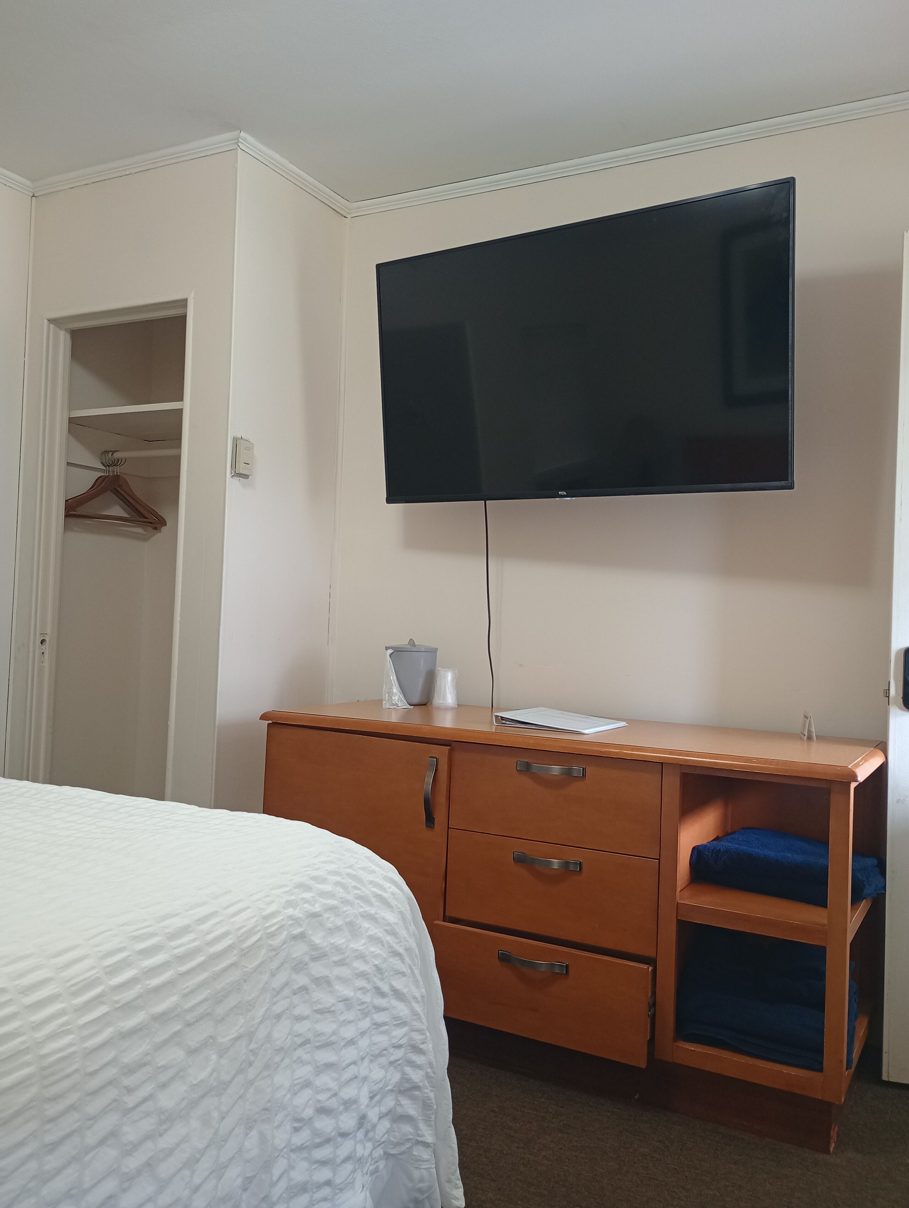 A hotel room with a bed, a wooden dresser with drawers, a flat-screen TV mounted on the wall, and a closet with a hanging garment.