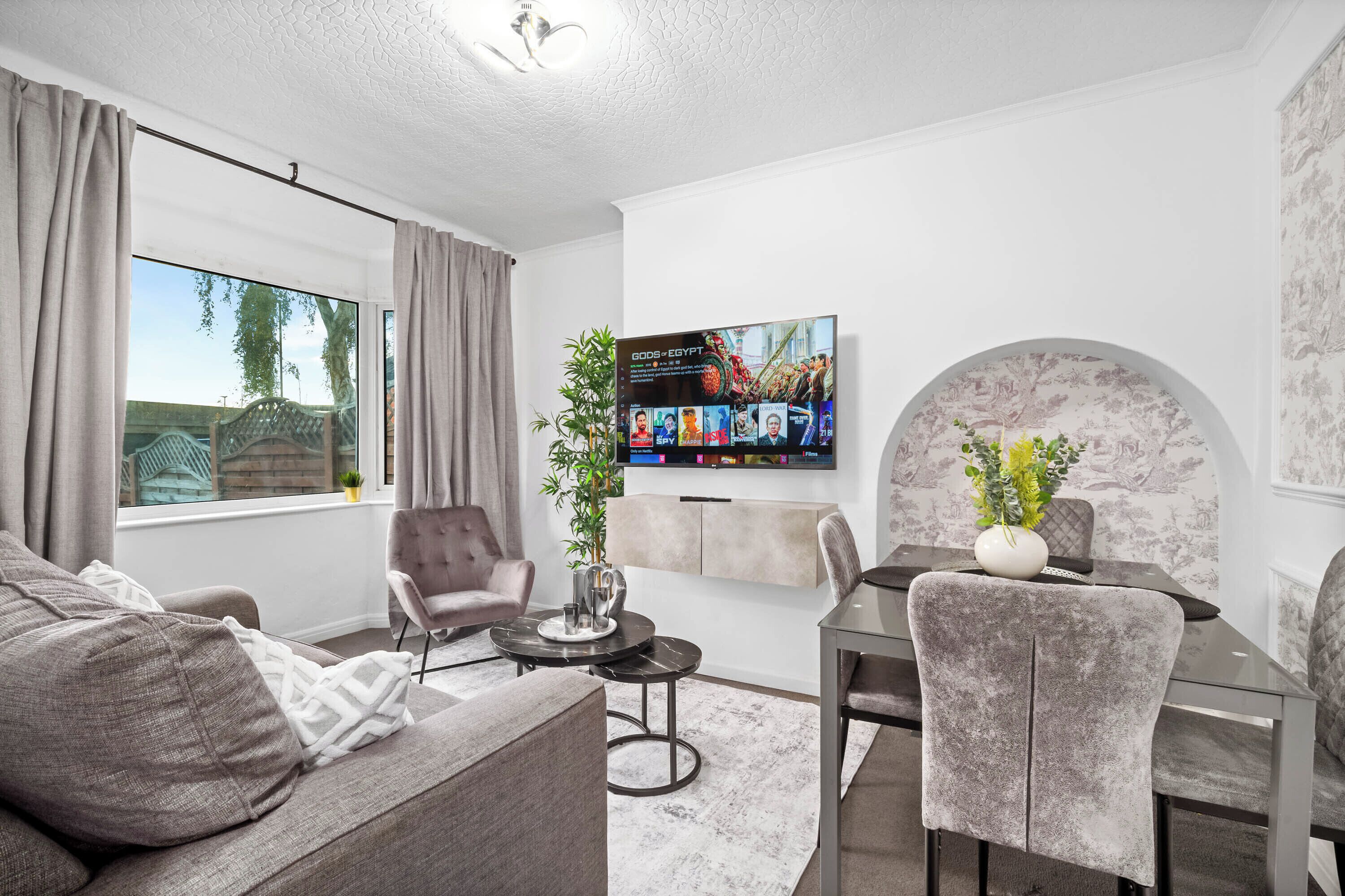 A modern living room with a sofa, armchair, dining table, and a TV mounted on the wall.