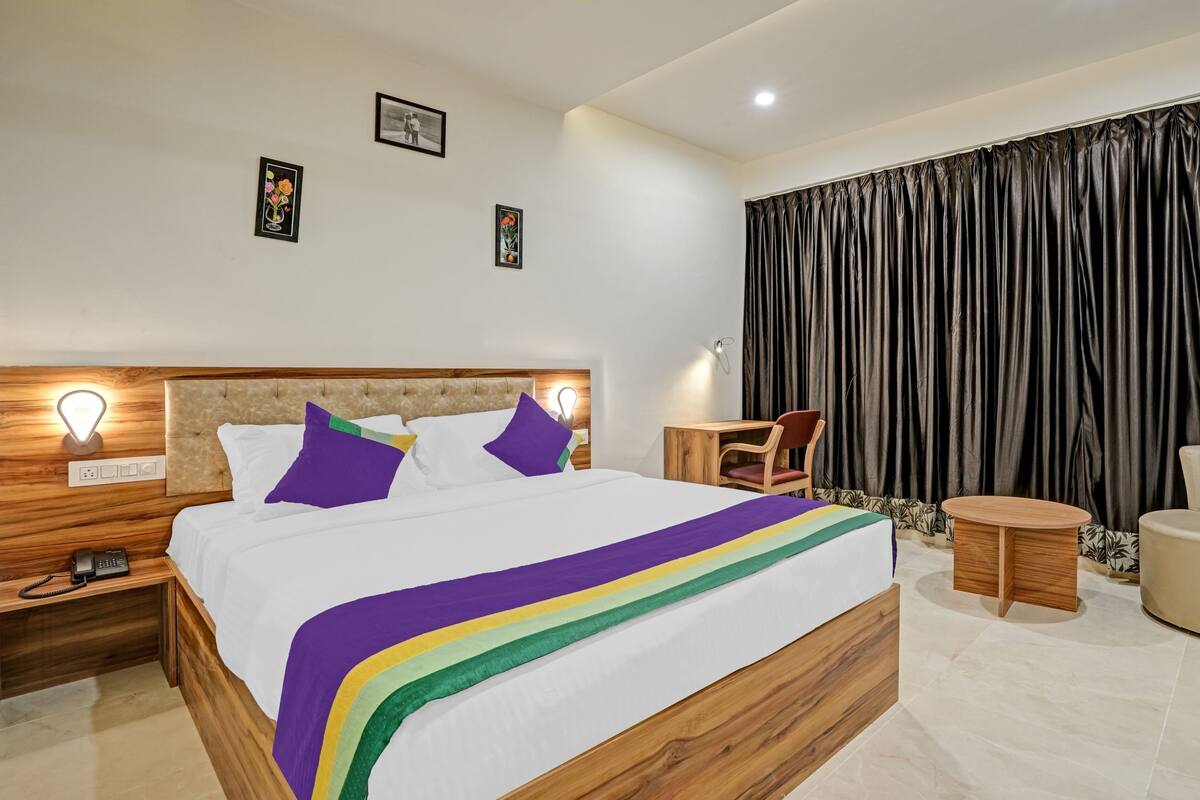 Quarto de hotel moderno com cabeceira de madeira, cama com roupa de cama branca e um cobertor listrado em tons de arco-íris, mesa de cabeceira de madeira com um telefone, uma pequena mesa de madeira, uma poltrona bege e uma janela grande com cortinas pretas.