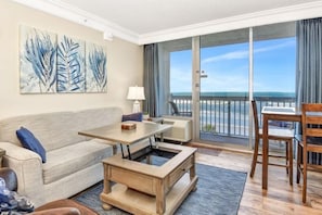 A modern living room with a sofa, a coffee table, a dining table with chairs, and a large sliding glass door leading to a balcony with a view of the ocean.