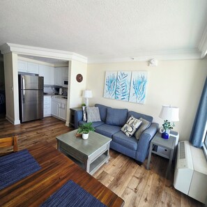 A living room with a sofa, coffee table, and a small side table with a lamp. There is a kitchen visible in the background. - Gorgeous 1 bdrm private balcony, kitchen direct oceanfront, pool hot tub, sauna (Daytona Beach)