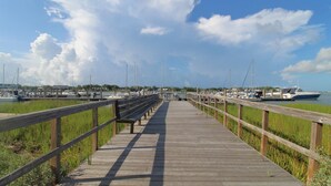 A wooden pier leading to a marina with boats docked, surrounded by grassy areas and a clear sky. - Lovely Long Term Rental~ Marsh Nest (Folly Beach)