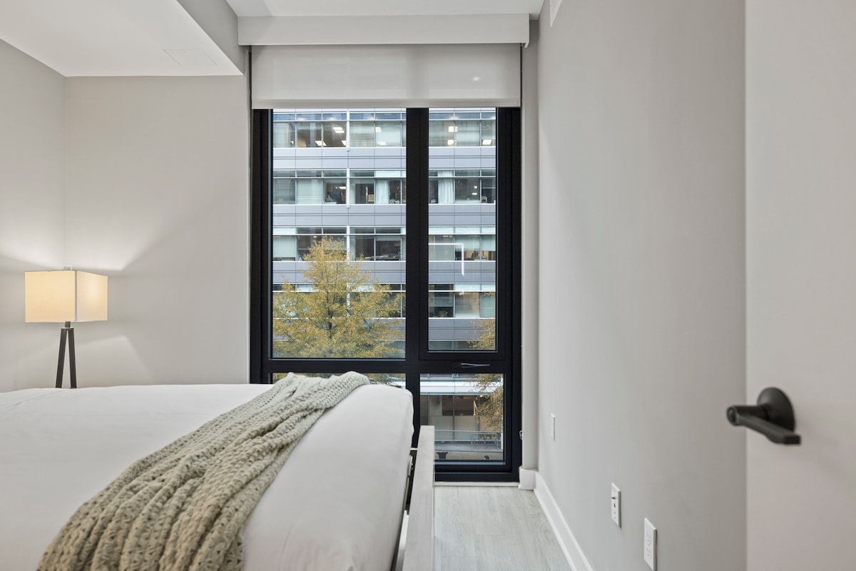 A modern hotel room with a large bed, a bedside lamp, a window with a view of a building and trees, and a door with a handle.