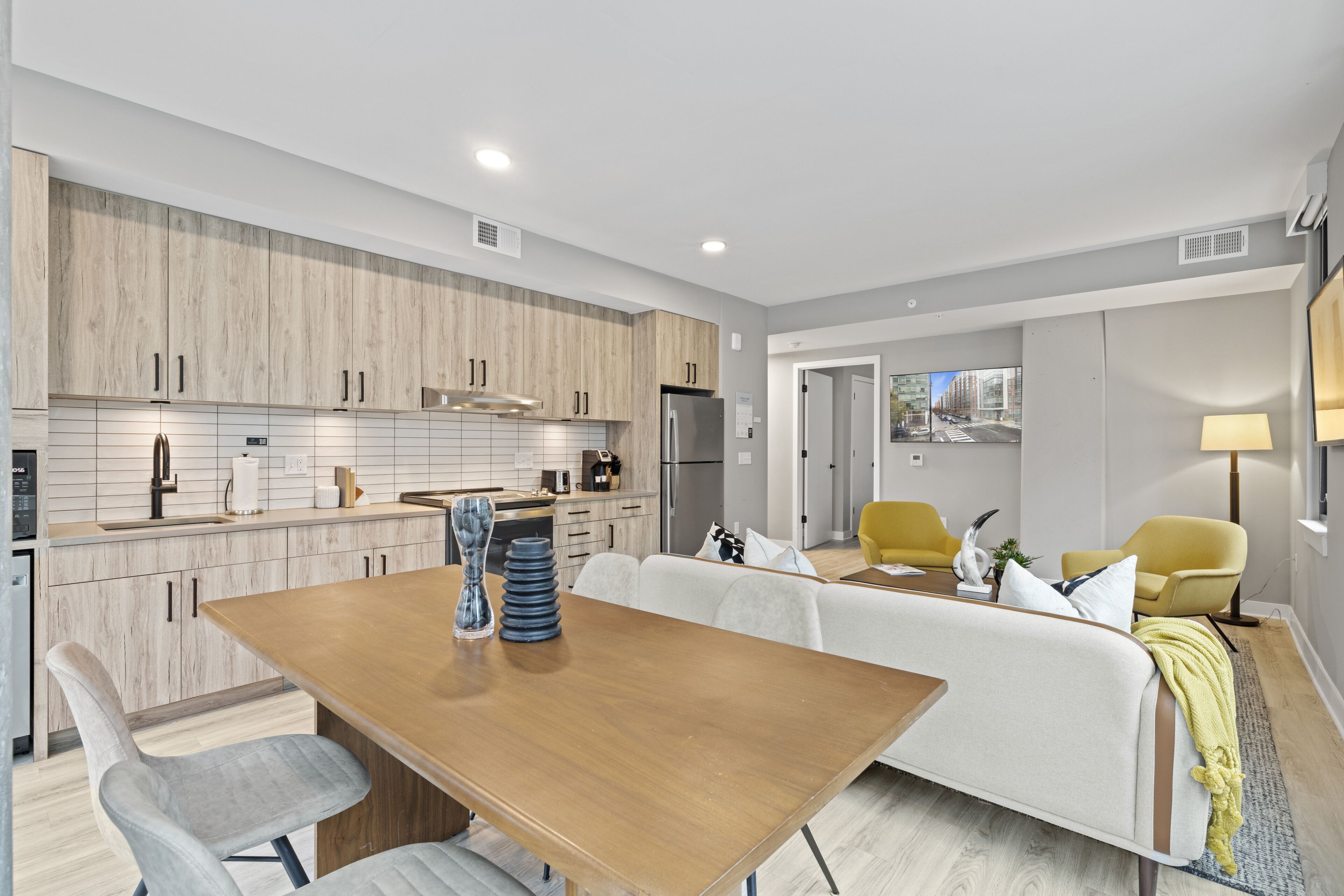A modern kitchen with wooden cabinets, a dining area with a wooden table, and a living room with a sofa and yellow chairs.