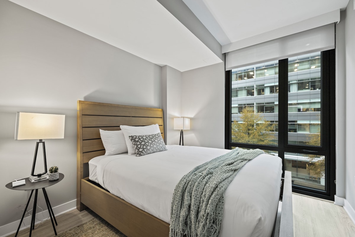 A modern hotel room with a large bed, a bedside table with a lamp, and a view of a building through a large window.