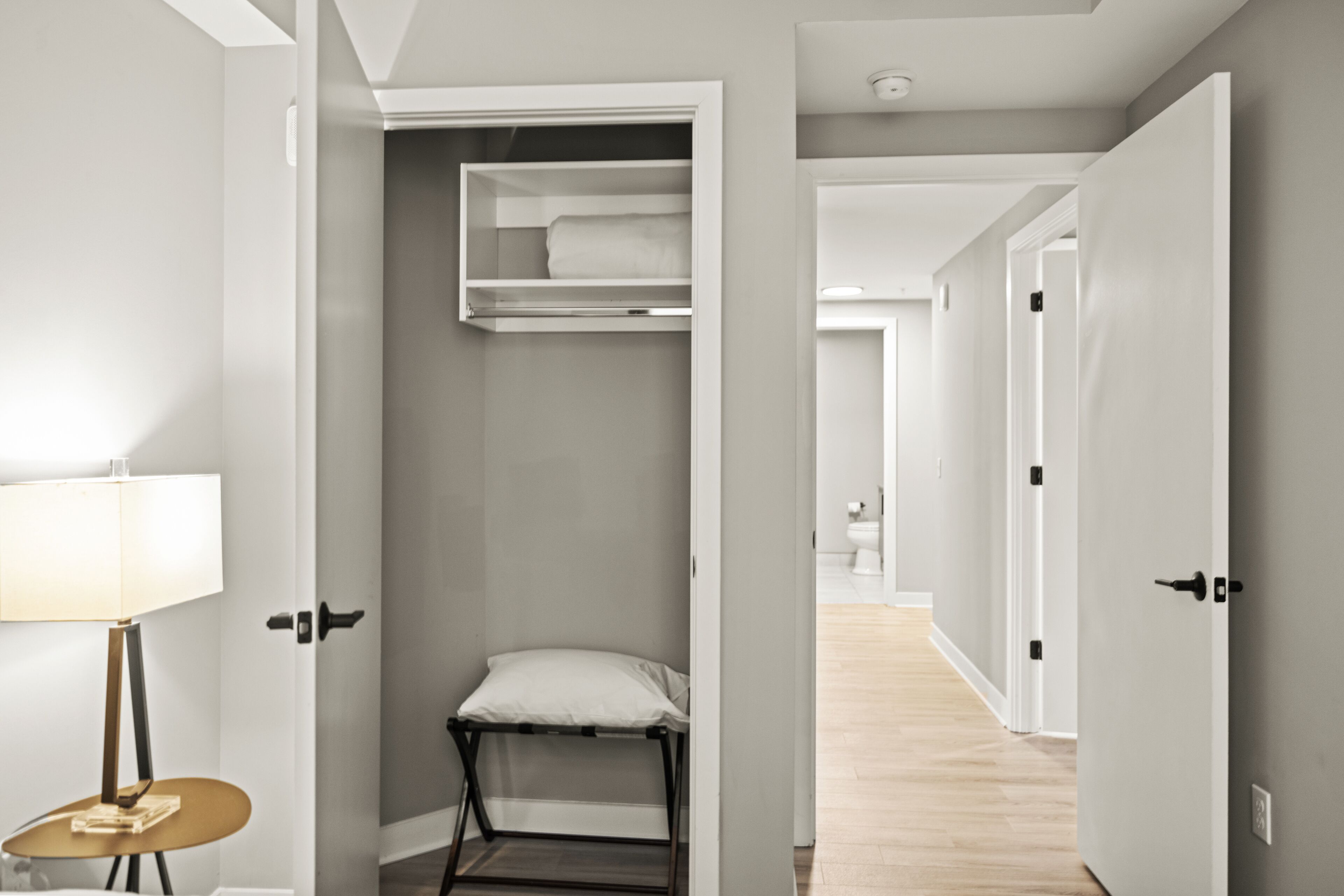A hallway with a bed in the background, a chair, and a lamp.
