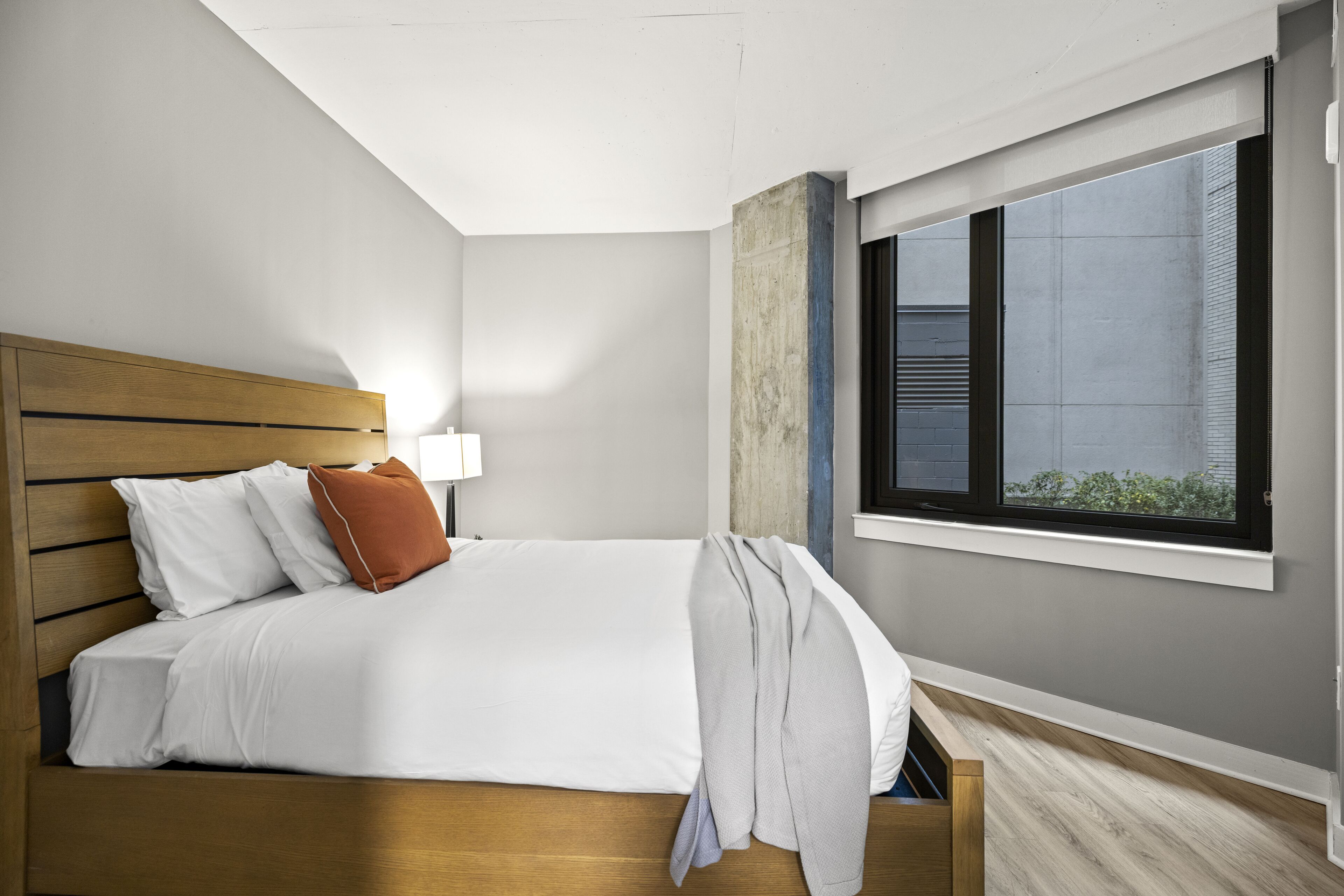 A modern bedroom with a wooden headboard, white bedding, a grey throw blanket, and a window with a view of greenery.